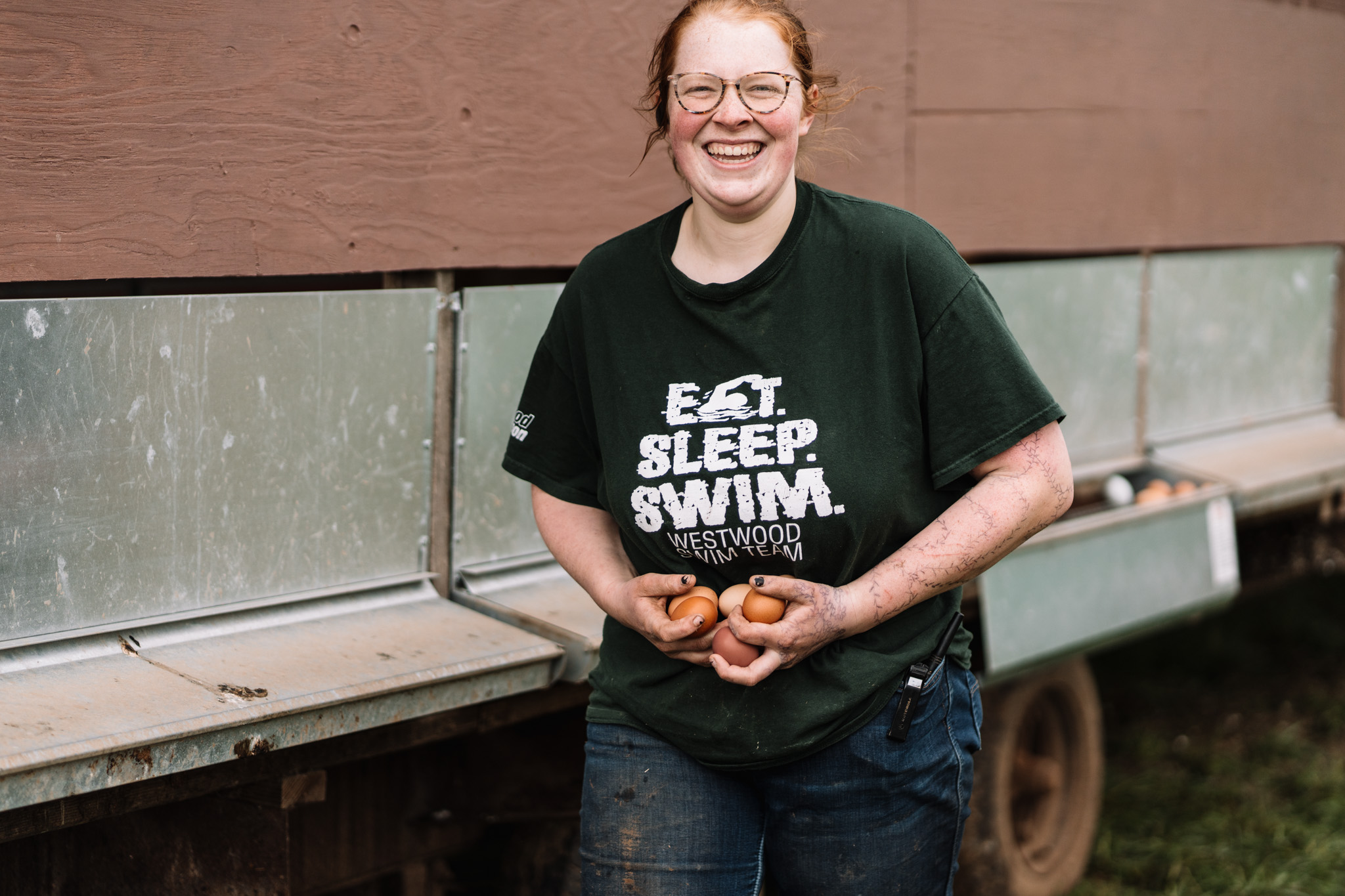 Farm worker smiling while pouring feed into a bin beside pasture infrastructure.